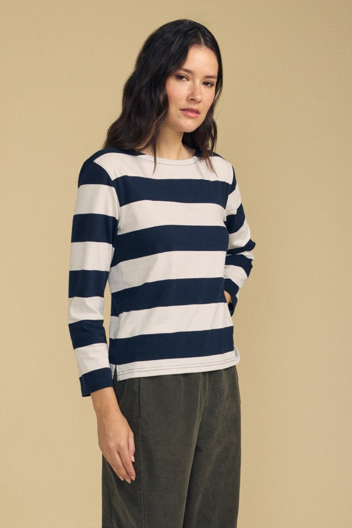 A woman stands against a beige background wearing the GABRIELA TEE by Velvet by Graham & Spencer, a relaxed fit, long sleeve striped cotton jersey top in black and white, paired with olive green pants.-41860456284353