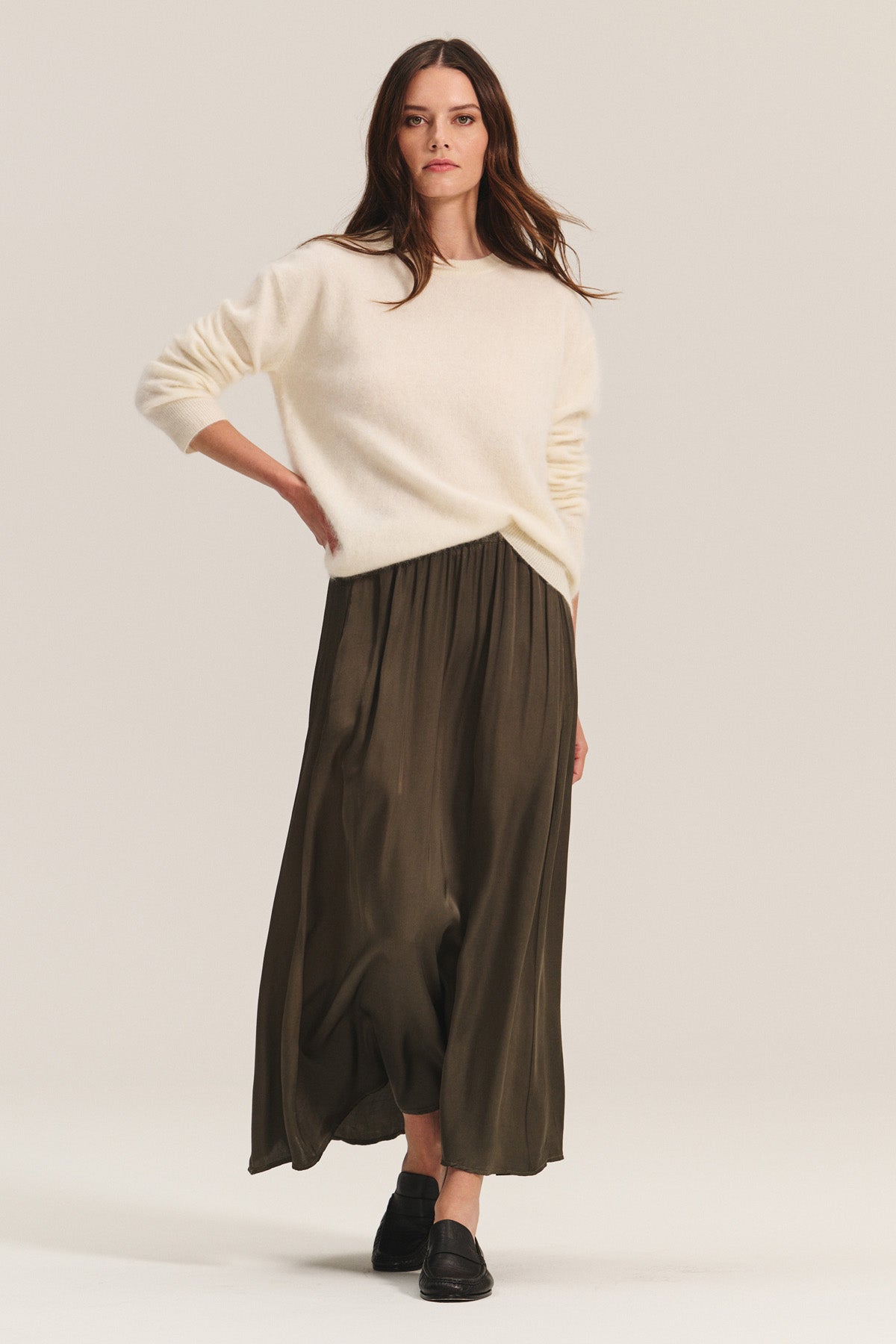 A woman poses against a plain light background wearing a cream sweater, black loafers, and the JUNIPER SKIRT by Velvet by Graham & Spencer—an elegant olive green satin skirt with an elastic waistband.-40263650377921