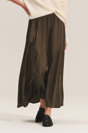 A person wears the JUNIPER SKIRT by Velvet by Graham & Spencer—an elegant olive green satin skirt with an elastic waistband—paired with a cream sweater and black loafers, photographed from the back against a neutral background.