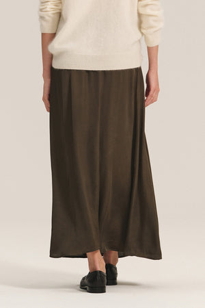 A person stands with their back to the camera, wearing Velvet by Graham & Spencer’s JUNIPER SKIRT in dark olive satin with an elastic waistband, a cream sweater, and black shoes against a plain background.
