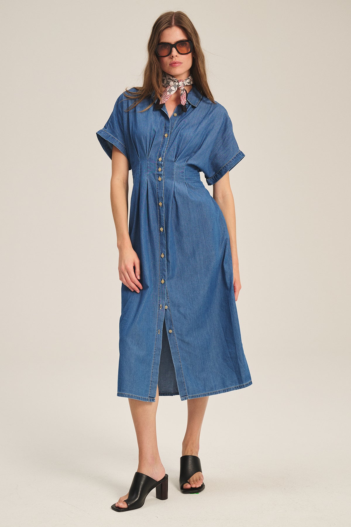 A woman wears the KARINA CHAMBRAY DRESS by Velvet by Graham & Spencer, styled with black heeled sandals, dark sunglasses, and a patterned neck scarf.-39600464429249