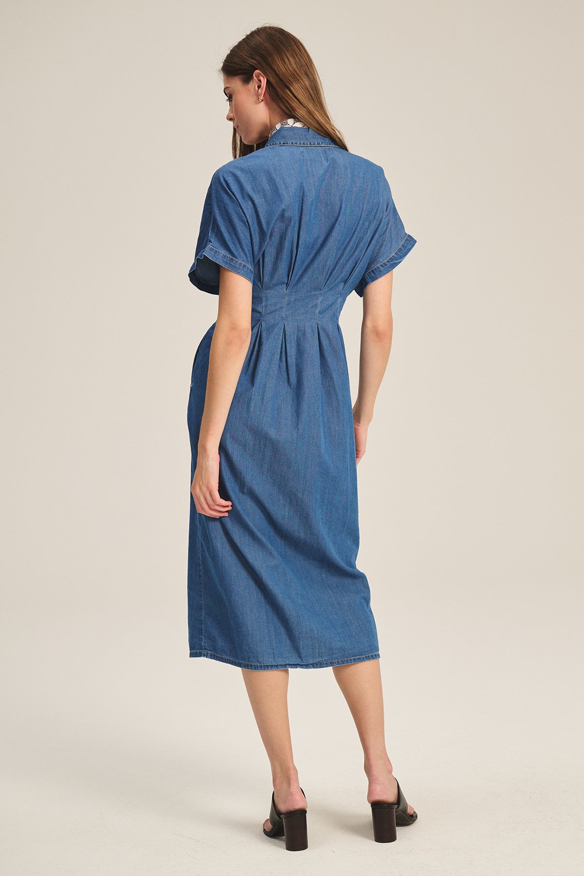 A woman stands with her back to the camera, wearing the KARINA CHAMBRAY DRESS by Velvet by Graham & Spencer and black heeled sandals against a plain background.