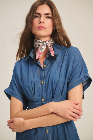 A woman in the KARINA CHAMBRAY DRESS by Velvet by Graham & Spencer, featuring short dolman sleeves, stands with arms crossed against a plain background, accessorized with a patterned scarf tied at her neck.