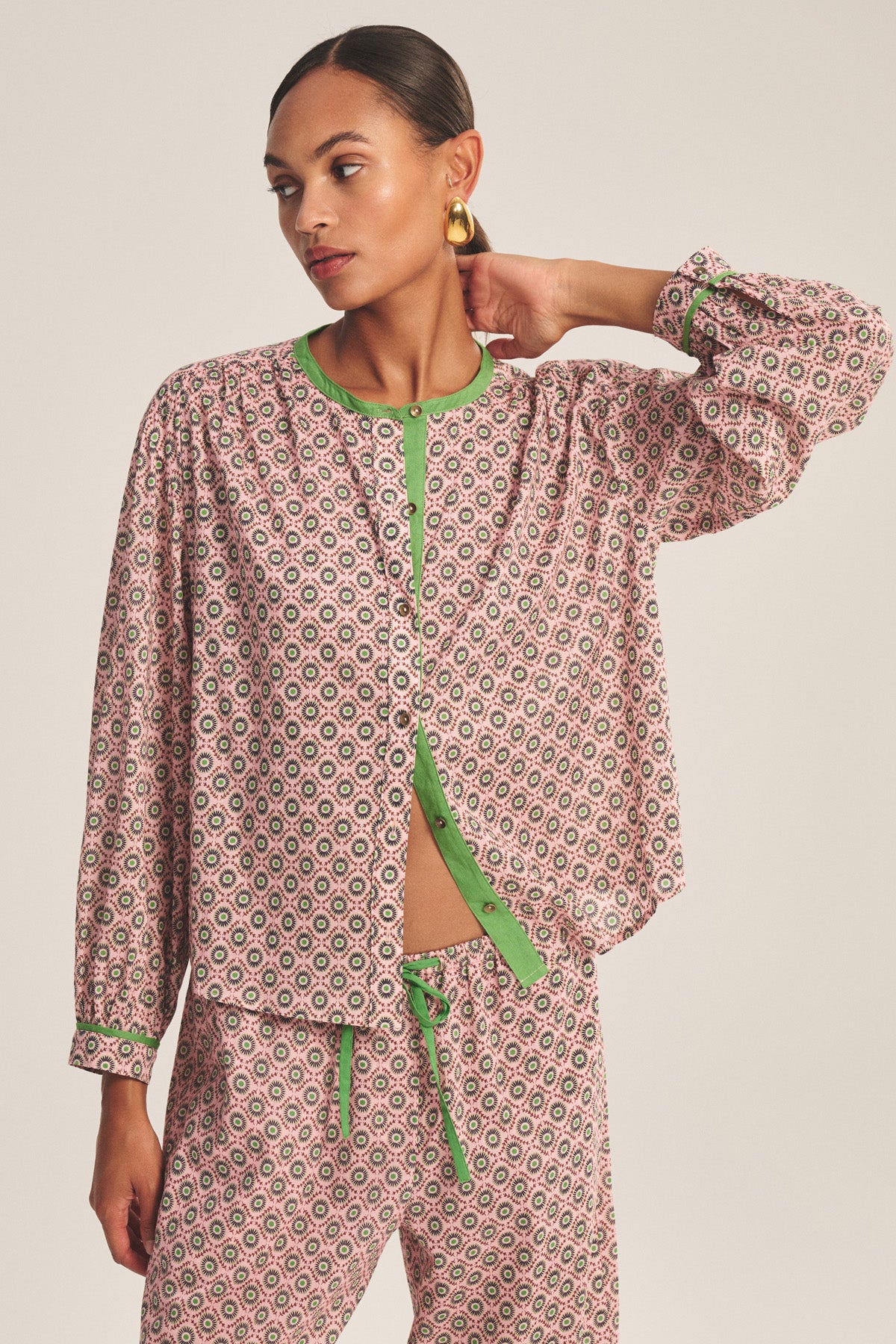 A person wearing the KARMEN TOP by Velvet by Graham & Spencer—a patterned pink and green button-front blouse—paired with matching drawstring pants, stands with one hand resting on their head.-41088948338881