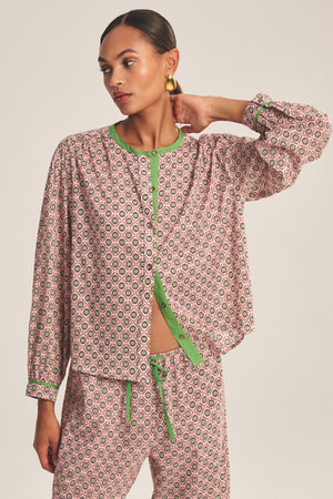 A person wearing the KARMEN TOP by Velvet by Graham & Spencer—a patterned pink and green button-front blouse—paired with matching drawstring pants, stands with one hand resting on their head.