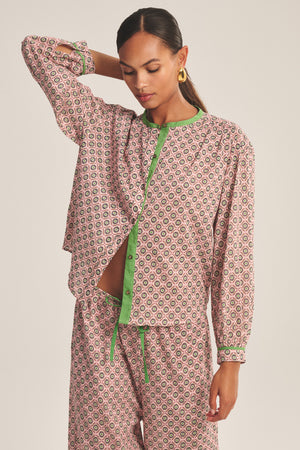 A woman wears the KARMEN TOP by Velvet by Graham & Spencer—a patterned pink and green pajama blouse with a relaxed fit and button-front closure, partially unbuttoned—posing against a light background.