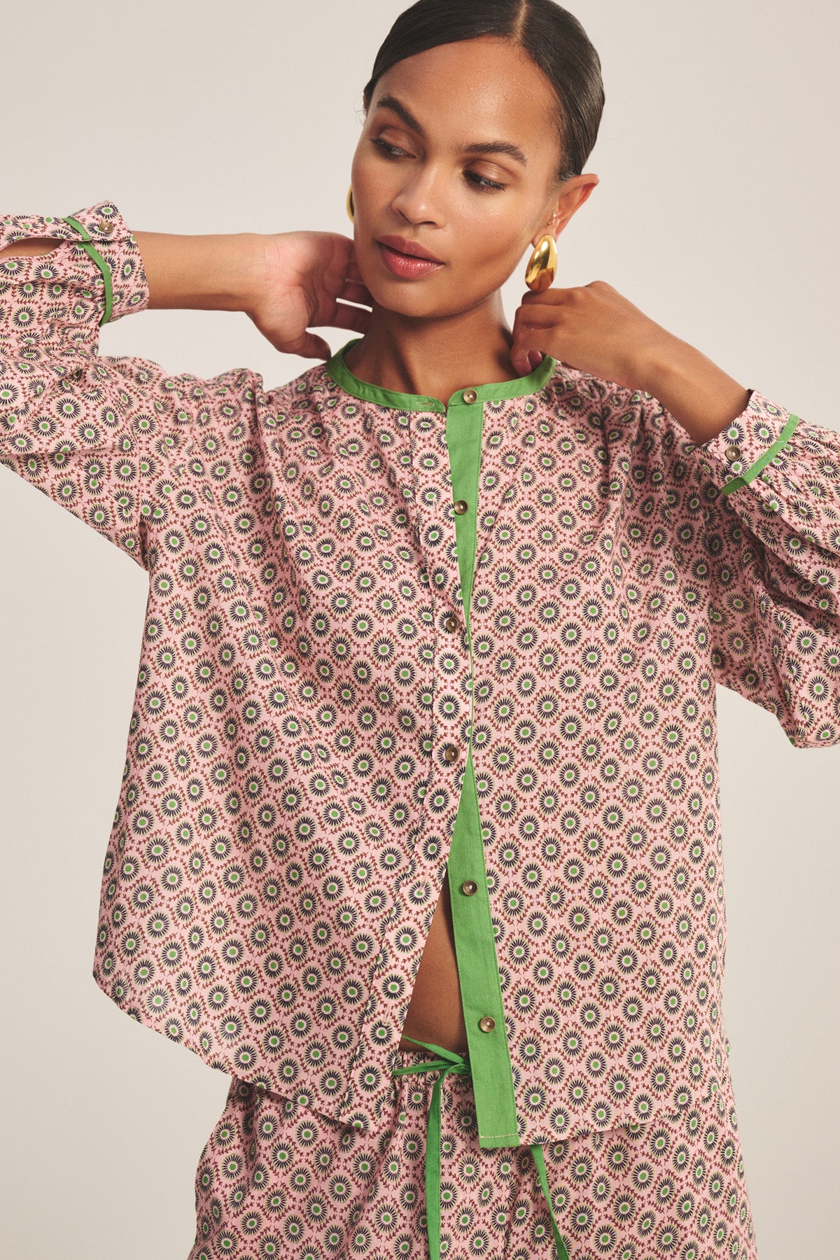 Woman wearing the Velvet by Graham & Spencer KARMEN TOP, a relaxed fit blouse with a patterned design and green trim, standing with hands at her neck, looking slightly downward.
