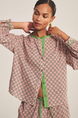 Woman wearing the Velvet by Graham & Spencer KARMEN TOP, a relaxed fit blouse with a patterned design and green trim, standing with hands at her neck, looking slightly downward.