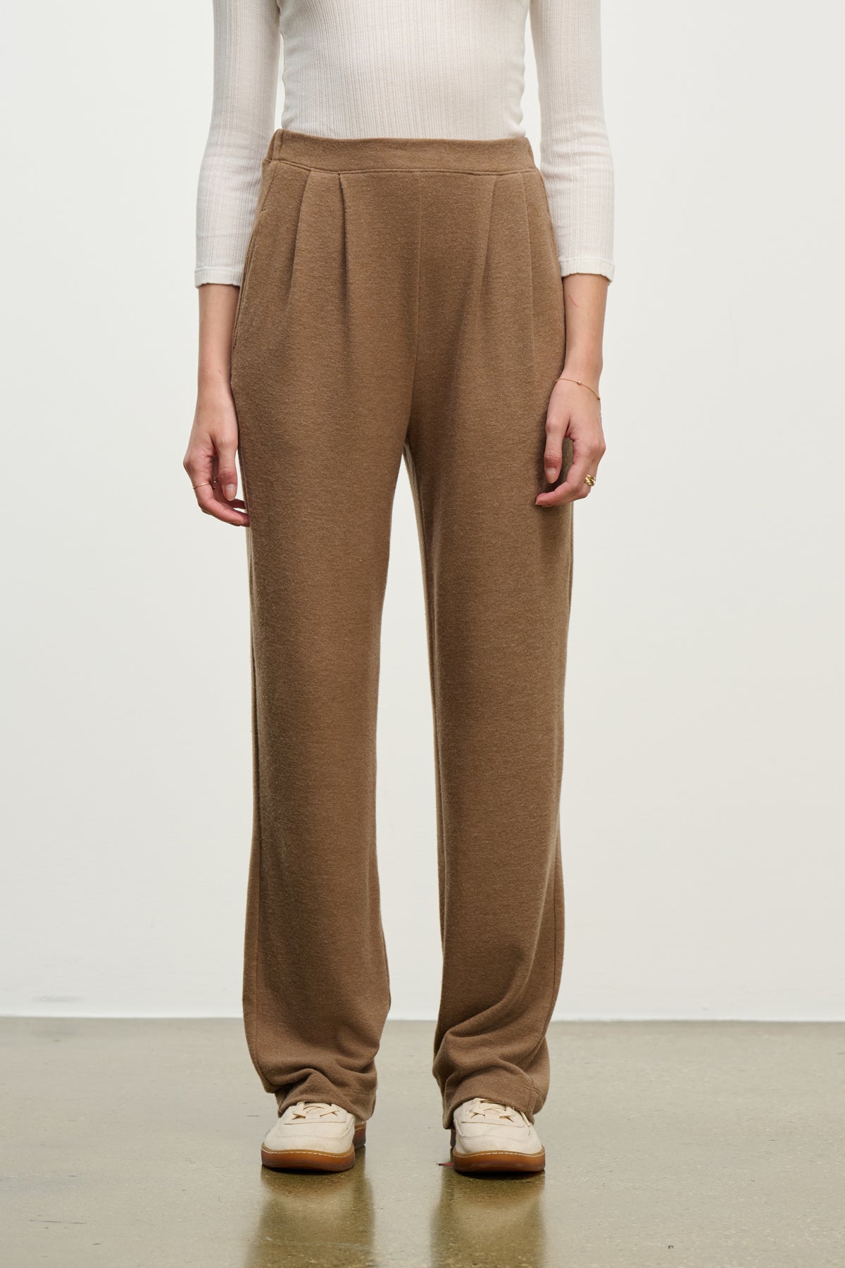 A person wears Velvet by Graham & Spencer's ELIYA SWEATPANT, a brown high-waisted lounge pant, paired with a white long-sleeve ribbed top and beige shoes, standing against a plain background.-39838349459649
