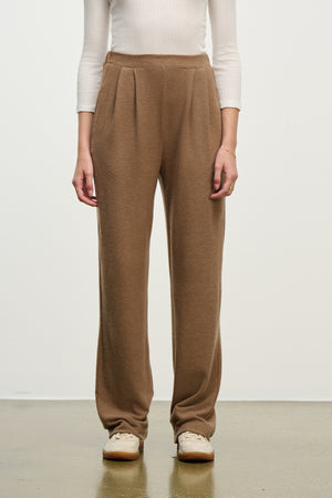 A person wears Velvet by Graham & Spencer's ELIYA SWEATPANT, a brown high-waisted lounge pant, paired with a white long-sleeve ribbed top and beige shoes, standing against a plain background.