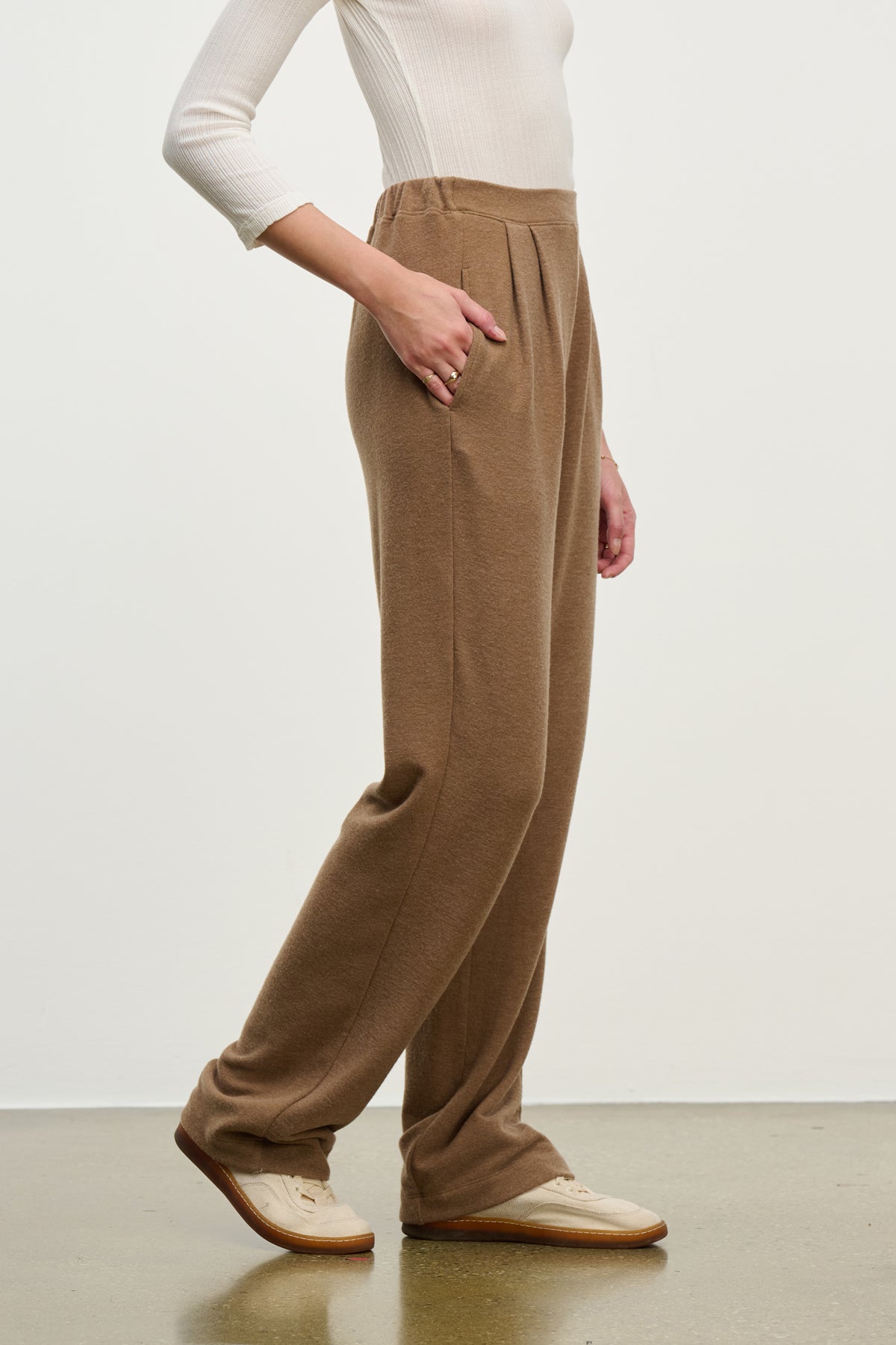   A person in a white long-sleeve shirt, ELIYA SWEATPANT by Velvet by Graham & Spencer—ultra-soft with pockets—and beige shoes stands on a light brown floor against a plain background. 