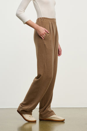 A person in a white long-sleeve shirt, ELIYA SWEATPANT by Velvet by Graham & Spencer—ultra-soft with pockets—and beige shoes stands on a light brown floor against a plain background.