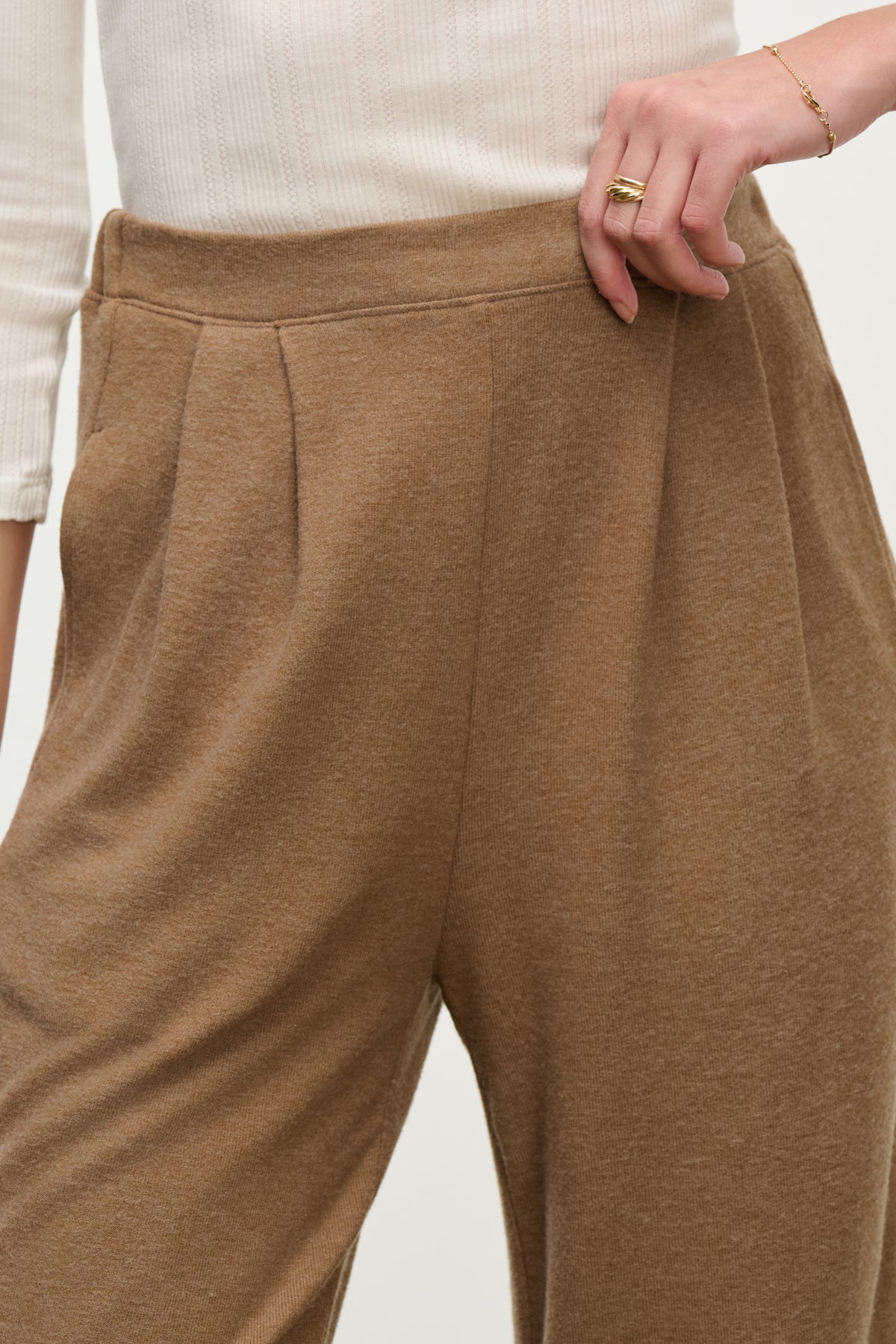   A person wearing ultra-soft, high-waisted ELIYA SWEATPANT in brown by Velvet by Graham & Spencer with a comfortable fit and a white ribbed top, one hand resting on the waistband. 