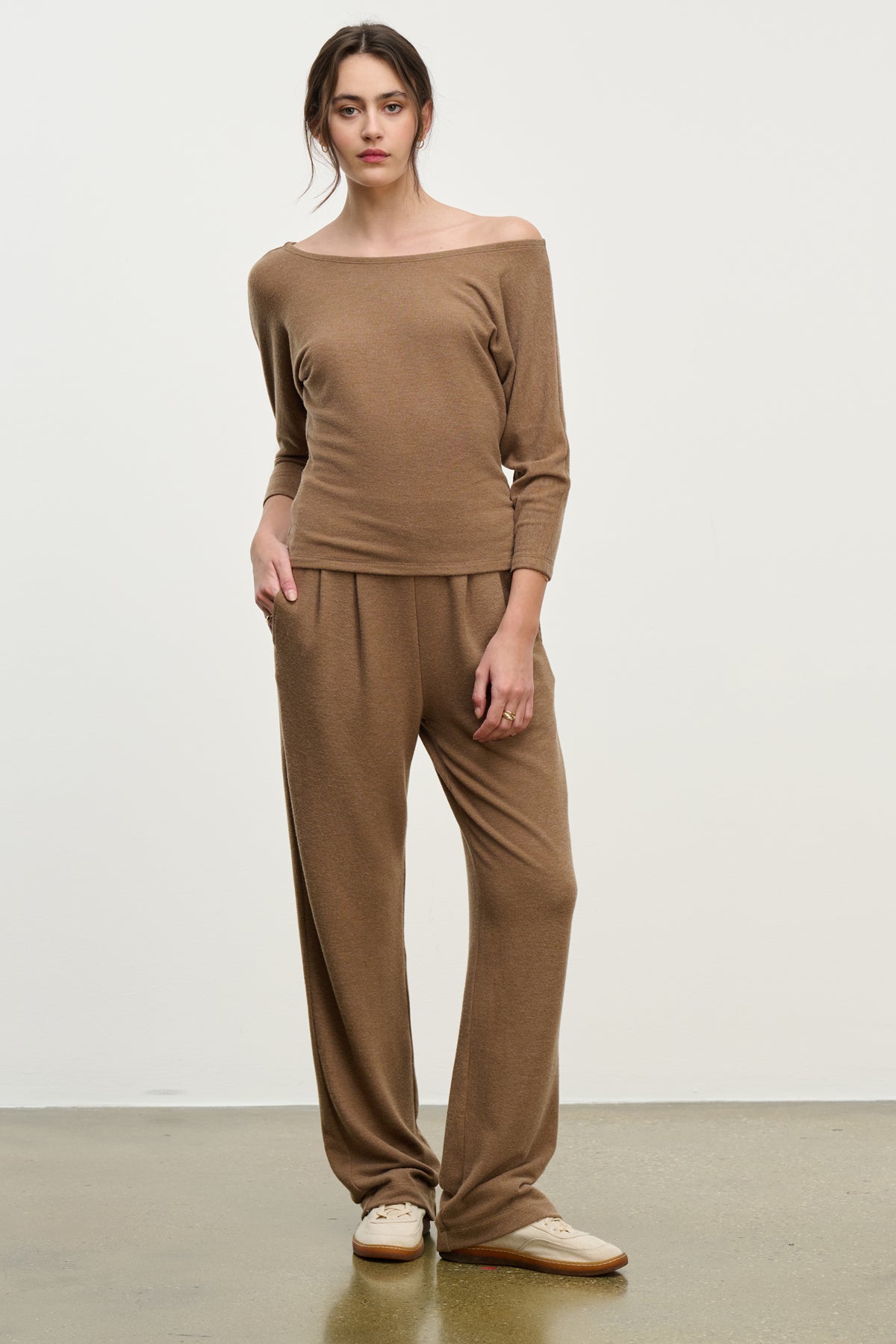 A woman stands against a plain background wearing Velvet by Graham & Spencer's ELIYA SWEATPANT—ultra-soft, wide-leg sweatpants with a comfortable fit—paired with a loose brown off-shoulder top and beige shoes.-39838349328577