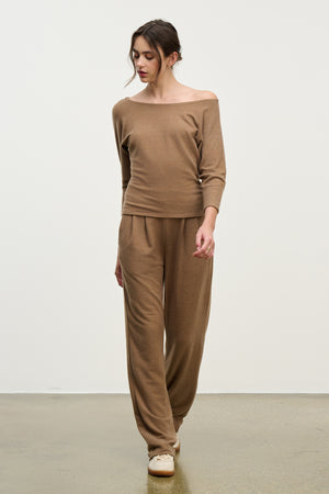 A woman stands indoors wearing Velvet by Graham & Spencer’s ELIYA SWEATPANT, an ultra-soft, light brown sweatpant with a comfortable fit, paired with a matching off-shoulder top and beige slip-on shoes against a plain background.