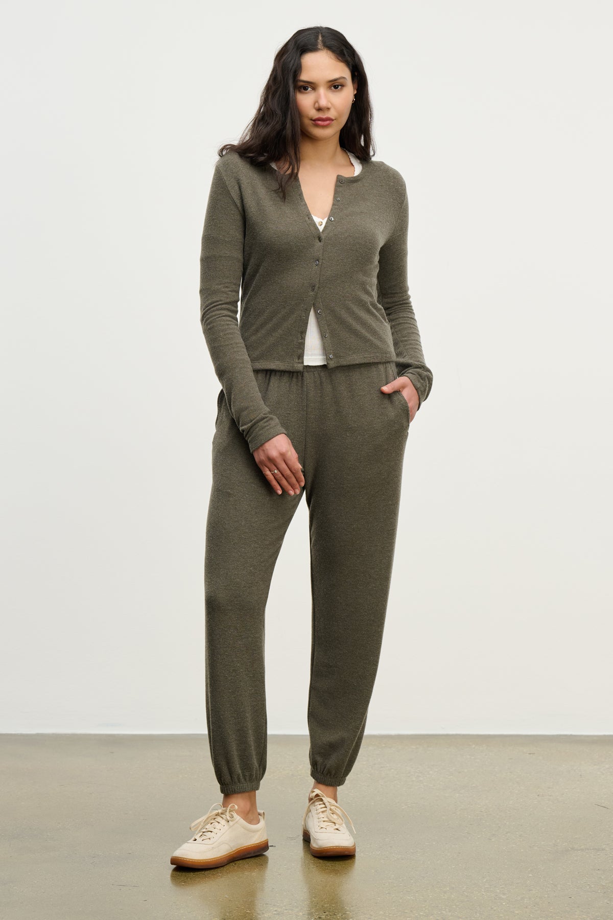   A woman stands against a plain background wearing Velvet by Graham & Spencer's olive green LEXIE SWEATPANT with an elastic waistband, paired with a matching cardigan and beige sneakers. 