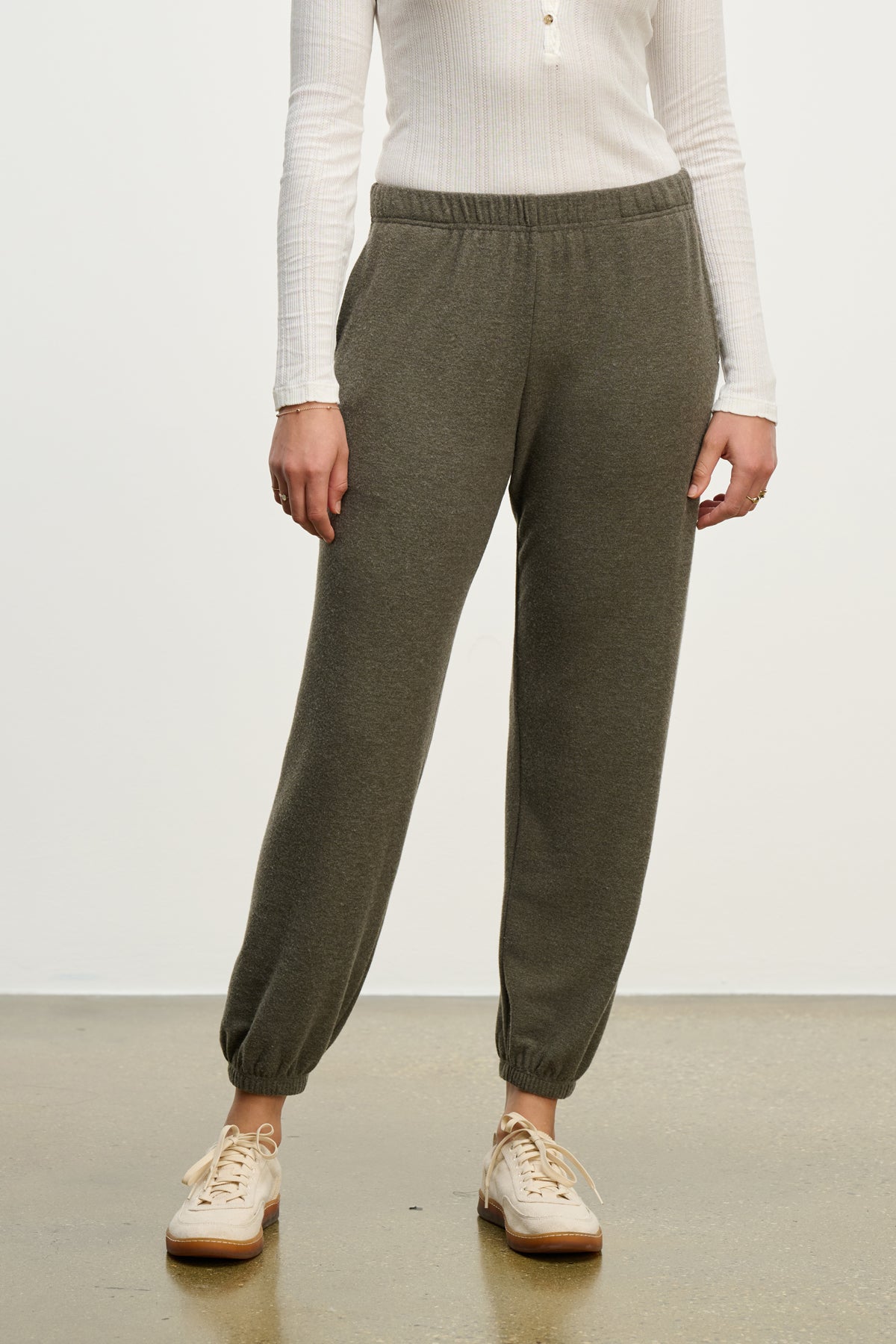   A person wears LEXIE SWEATPANT by Velvet by Graham & Spencer, featuring an olive green hue and elastic waistband, paired with a white ribbed long-sleeve shirt and beige sneakers, standing on a concrete floor against a light background. 