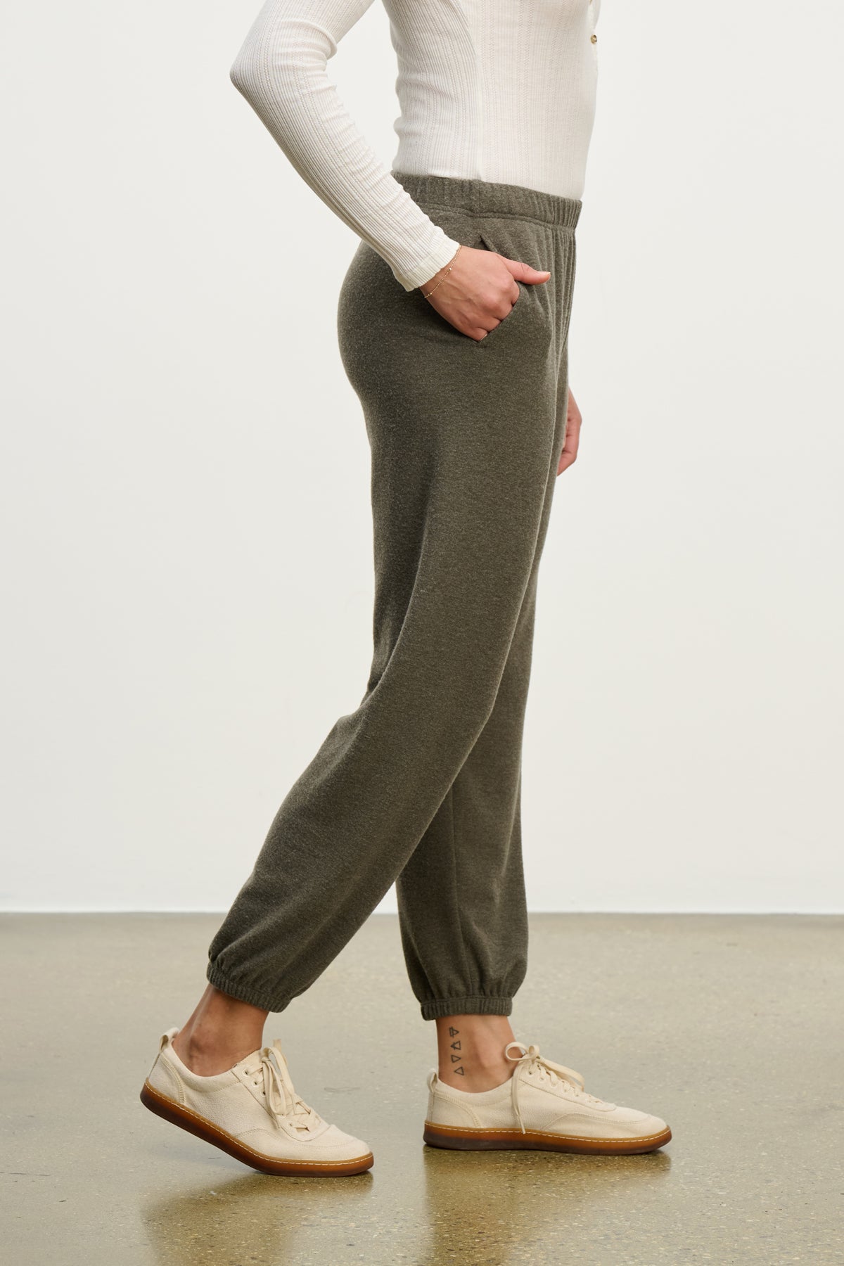 A person stands on a polished floor in a white long-sleeve top, Velvet by Graham & Spencer LEXIE SWEATPANT in gray with an elastic waistband, and beige lace-up shoes, shown from the side.-39838358077633