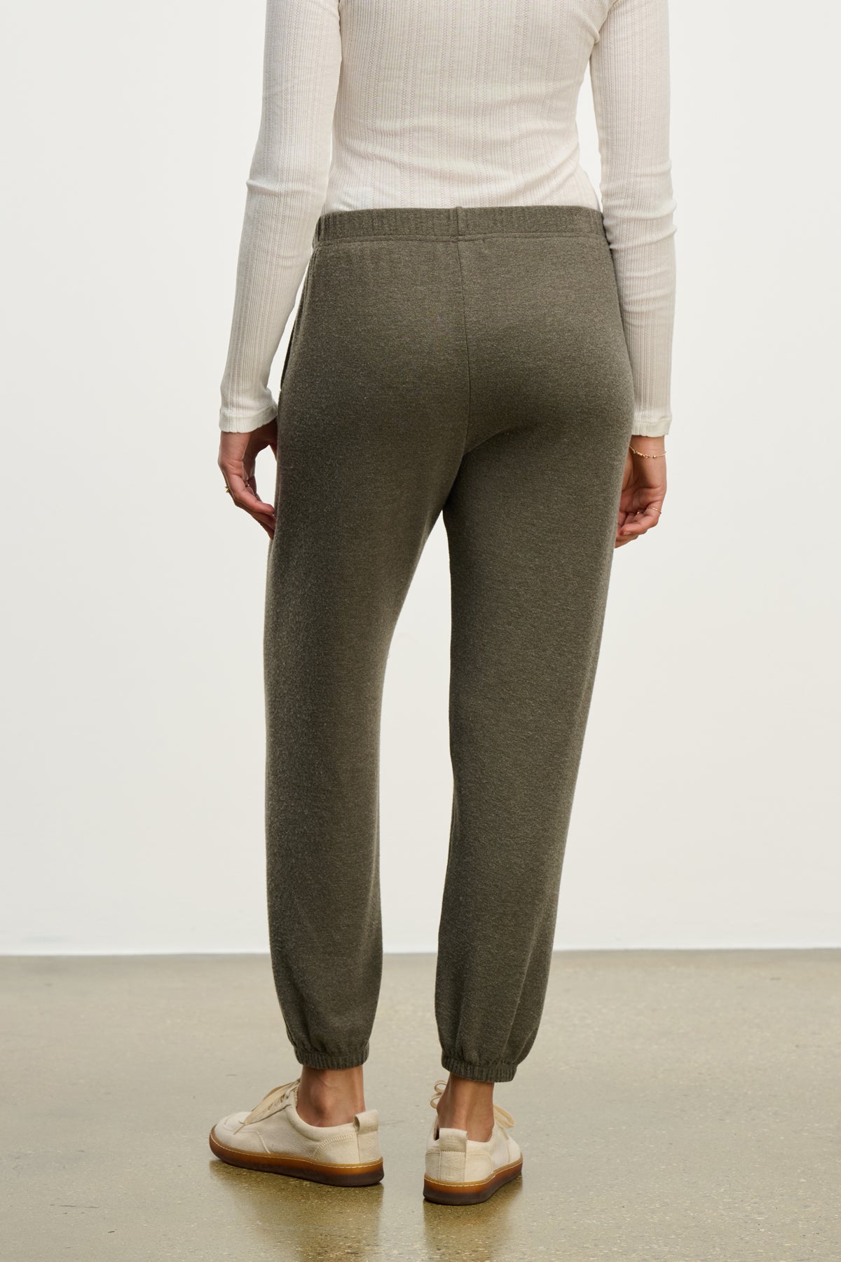 Person shown from the back wearing the LEXIE SWEATPANT by Velvet by Graham & Spencer—olive green joggers with an elastic waistband—paired with a long-sleeve white shirt, standing on a light brown floor.-39838358044865