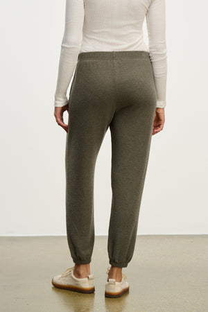Person shown from the back wearing the LEXIE SWEATPANT by Velvet by Graham & Spencer—olive green joggers with an elastic waistband—paired with a long-sleeve white shirt, standing on a light brown floor.