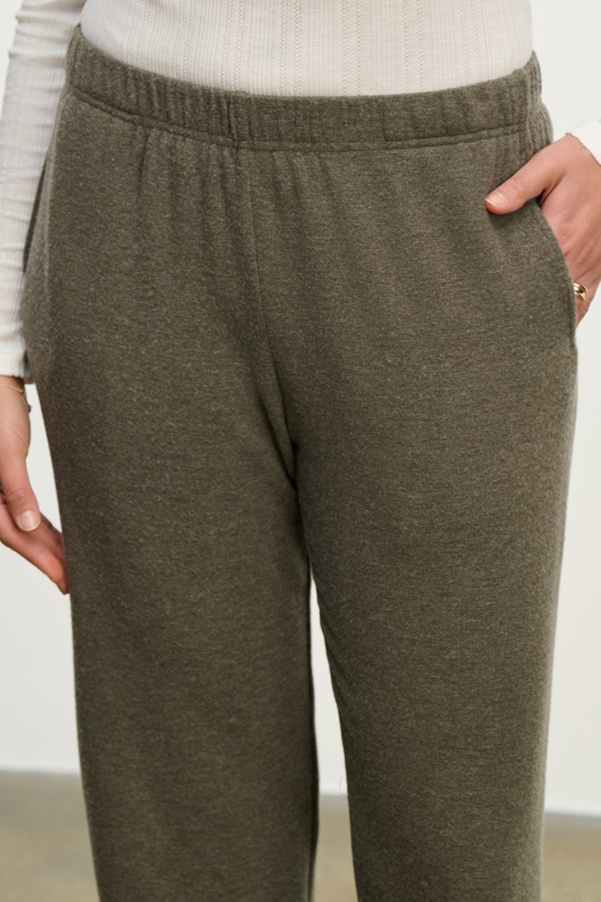   A person wearing the LEXIE SWEATPANT by Velvet by Graham & Spencer in dark gray, featuring a relaxed fit and elastic waistband, paired with a ribbed white long-sleeve top, hands tucked into the sweatpant pockets. 
