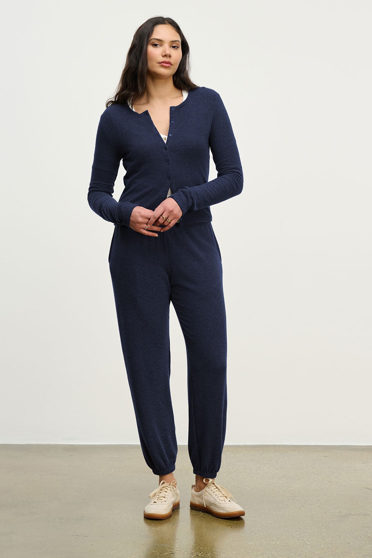 A woman stands indoors against a plain background, wearing the LEXIE SWEATPANT by Velvet by Graham & Spencer and beige sneakers.