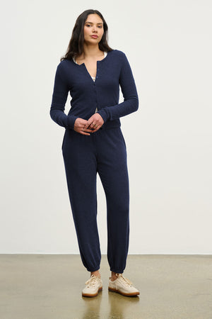 A woman stands indoors against a plain background, wearing the LEXIE SWEATPANT by Velvet by Graham & Spencer and beige sneakers.