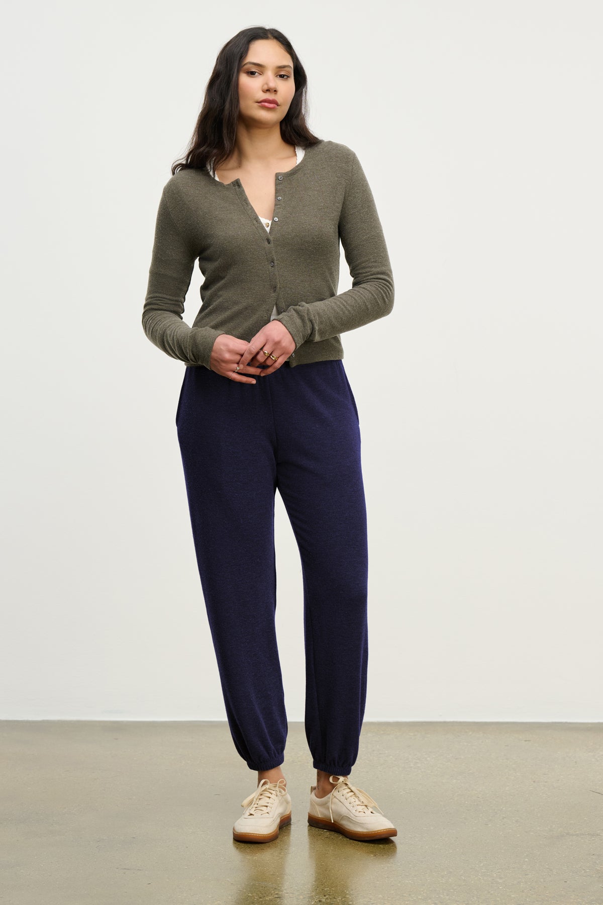   A woman stands against a plain background wearing an olive green buttoned cardigan, Velvet by Graham & Spencer's LEXIE SWEATPANT in navy with an elastic waistband, and beige sneakers. 
