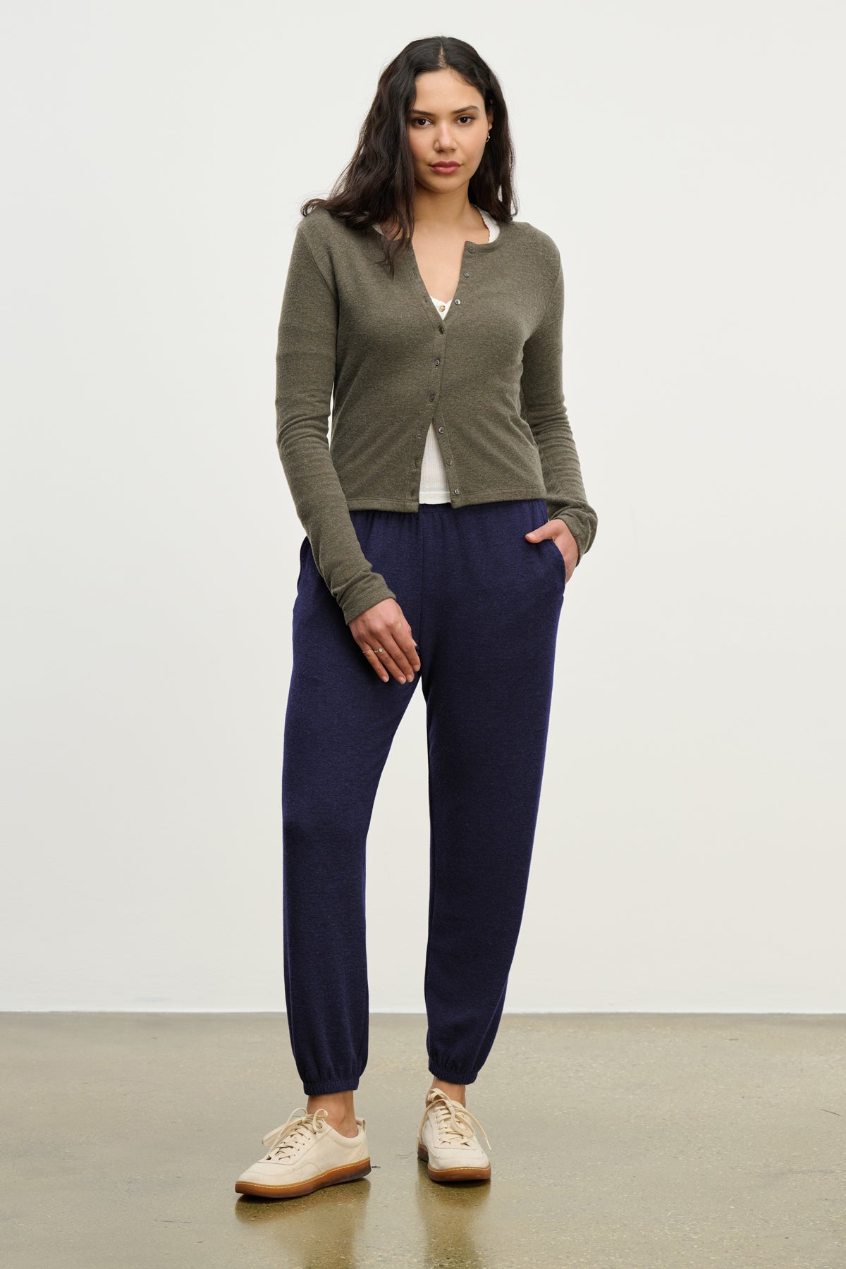  A woman stands against a plain background wearing Velvet by Graham & Spencer's LEXIE SWEATPANT in navy with an elastic waistband, paired with a khaki cardigan, white top, and beige sneakers, her hands tucked casually in her pockets. 