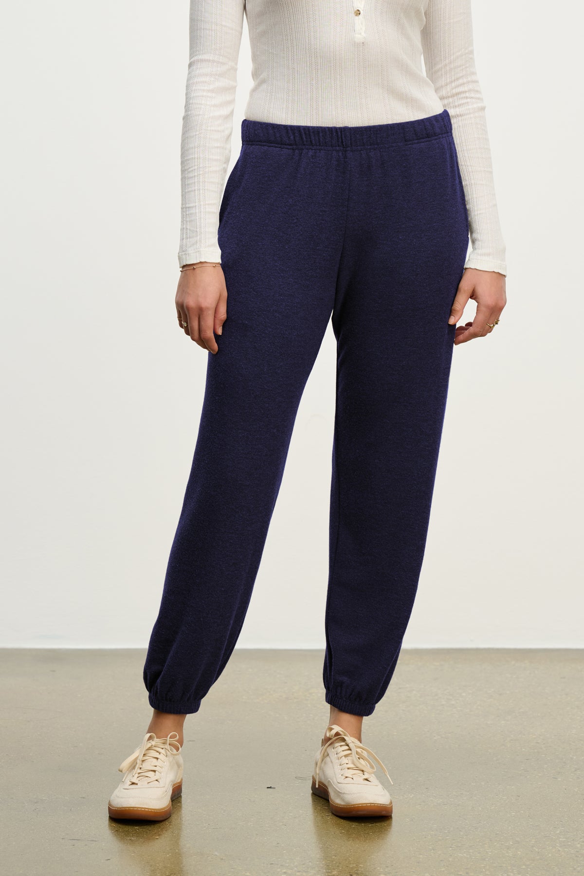   A person stands on a polished floor against a plain white background, wearing a white long-sleeve top, Velvet by Graham & Spencer's LEXIE SWEATPANT in dark blue with an elastic waistband, and beige sneakers. 