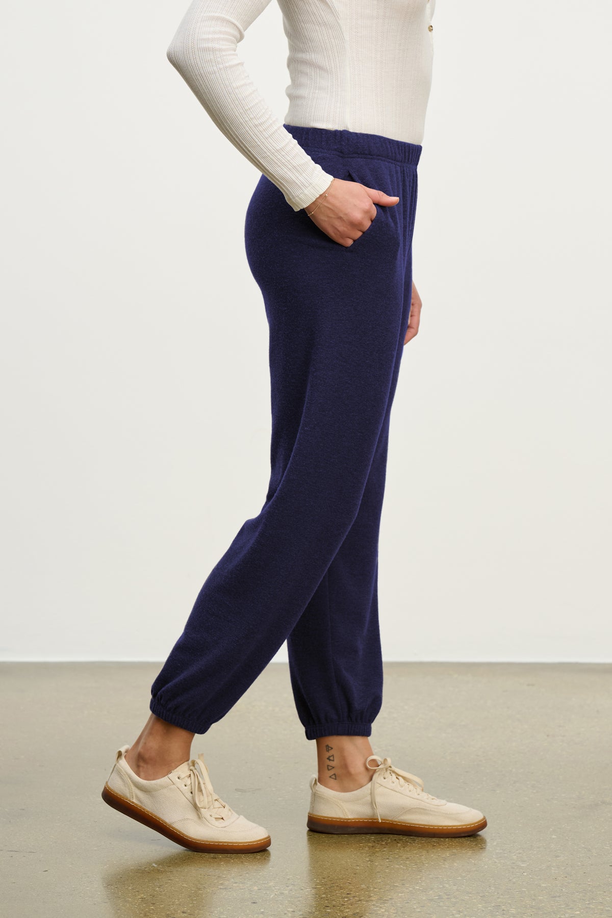  A person wearing a white long-sleeve top, Velvet by Graham & Spencer LEXIE SWEATPANT in navy with an elastic waistband, and beige lace-up shoes stands on a polished floor against a plain background. 
