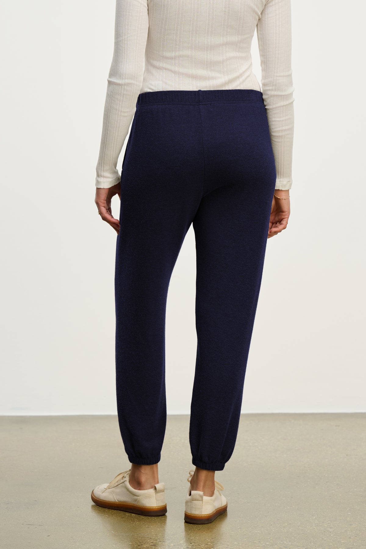   Model wearing the LEXIE SWEATPANT by Velvet by Graham & Spencer in navy blue, paired with a cream ribbed long-sleeve top and beige loafers, standing on a polished floor with their back to the camera. 