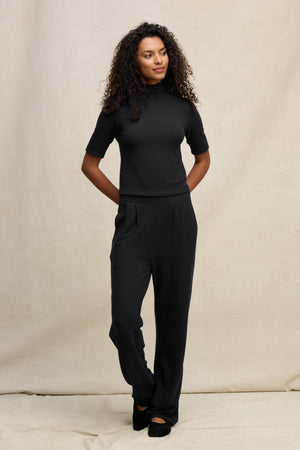 A woman with curly hair stands against a beige backdrop, wearing a black short-sleeve turtleneck and Velvet by Graham & Spencer's ELIYA SWEATPANT, an ultra-soft pair of sweatpants with a comfortable fit, her hands resting behind her back.