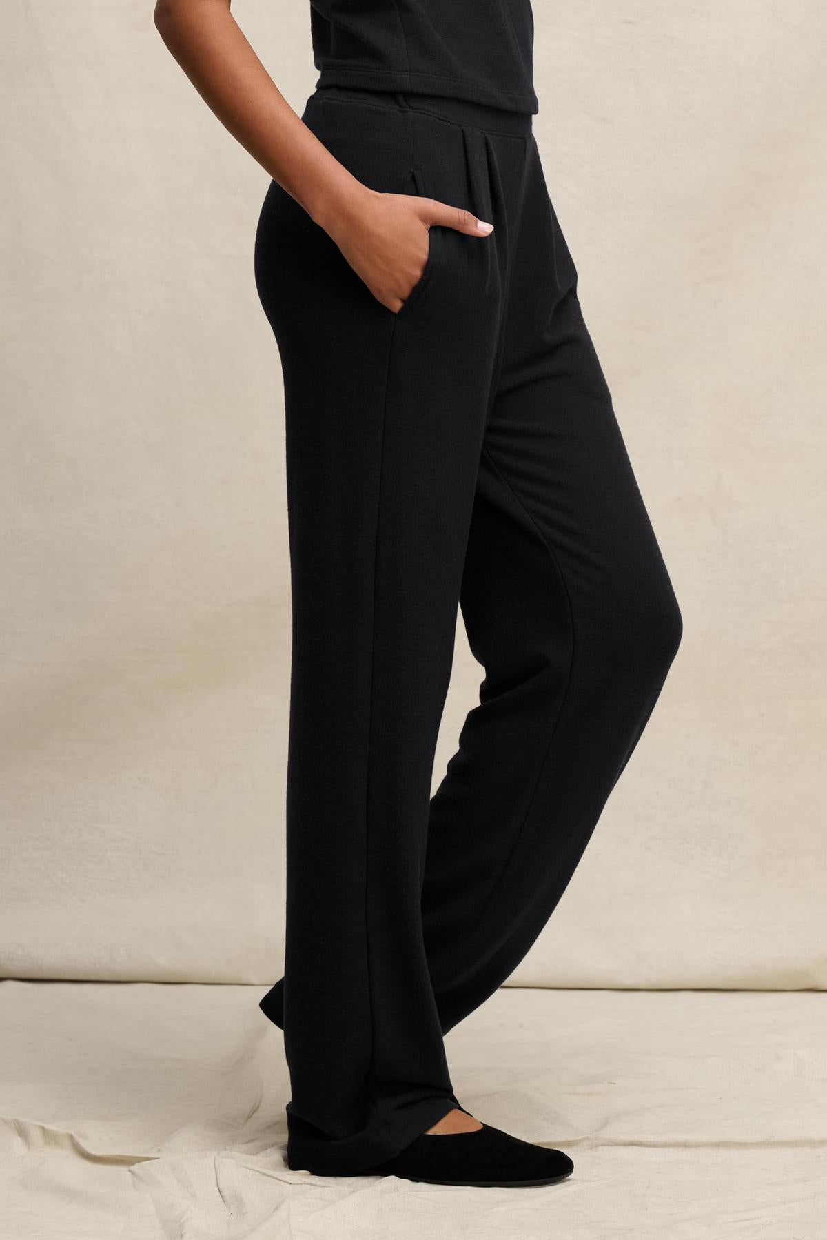 A person models the Velvet by Graham & Spencer ELIYA SWEATPANT—black, straight-leg sweatpants—paired with black shoes, standing with one hand in their pocket against a neutral background to showcase the comfortable fit.