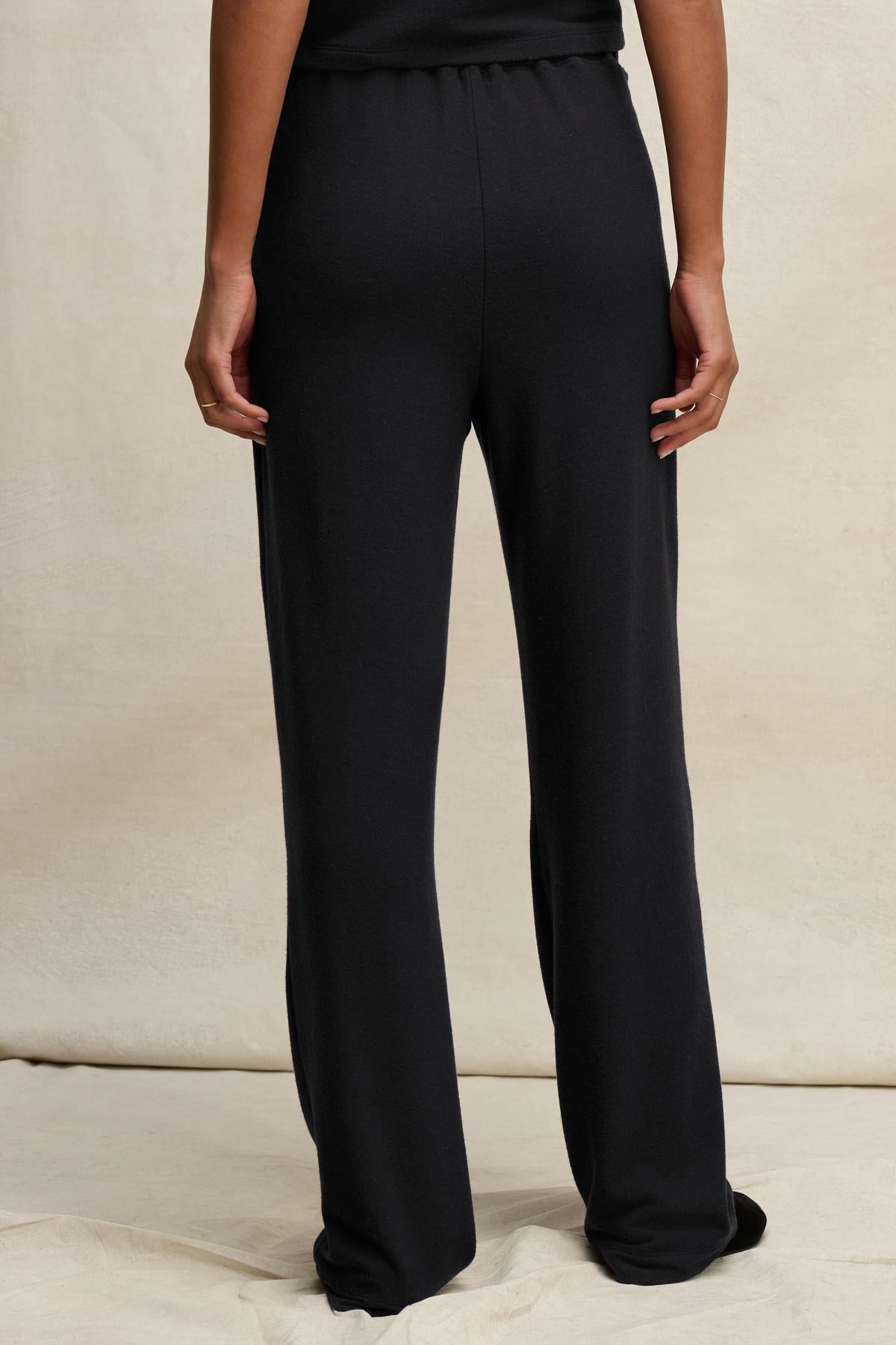A person is shown from the back, standing on a light surface and wearing the ELIYA SWEATPANT by Velvet by Graham & Spencer—black, straight-leg sweatpants paired with a matching black top for a comfortable fit.
