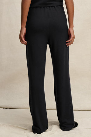 A person is shown from the back, standing on a light surface and wearing the ELIYA SWEATPANT by Velvet by Graham & Spencer—black, straight-leg sweatpants paired with a matching black top for a comfortable fit.