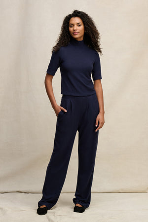 A woman stands against a neutral backdrop wearing Velvet by Graham & Spencer’s ELIYA SWEATPANT with a navy short-sleeve turtleneck top and black shoes.