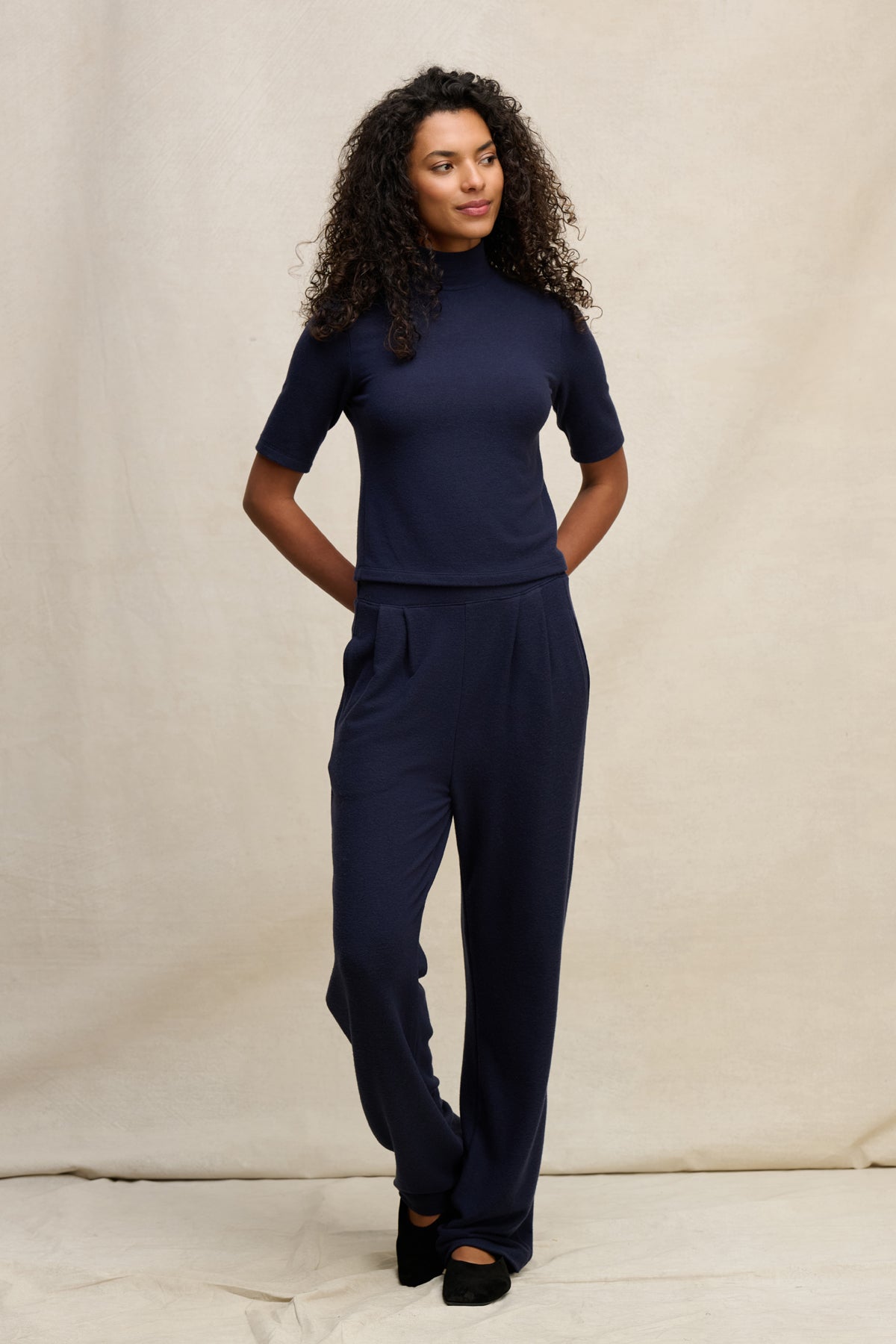 A woman stands against a neutral background wearing the Velvet by Graham & Spencer ELIYA SWEATPANT in an ultra-soft, straight-leg fit, paired with a navy short-sleeve top and her hands behind her back.-39838349230273