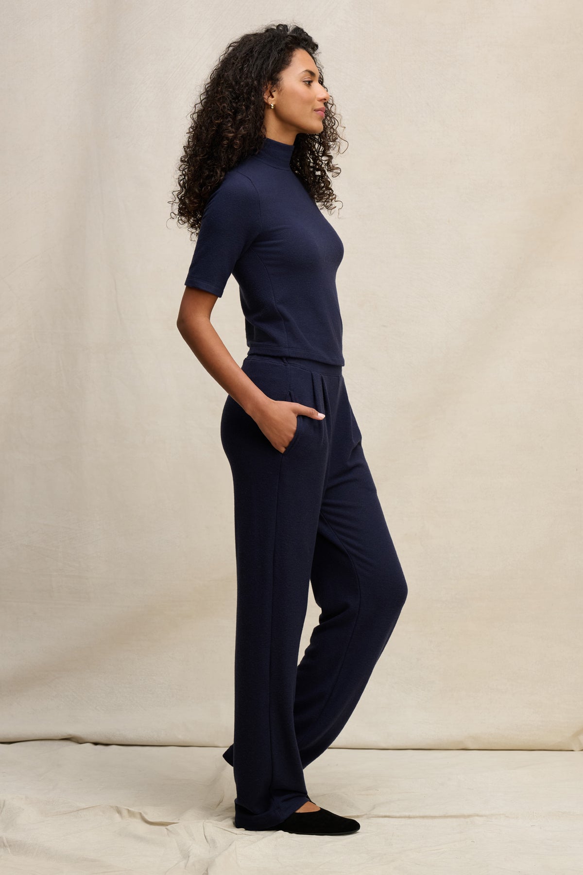 A woman with curly hair stands in profile, wearing a navy blue short-sleeve turtleneck and the Velvet by Graham & Spencer ELIYA SWEATPANT—ultra-soft, straight leg sweatpants with her hands in her pockets against a neutral background.-39838349197505