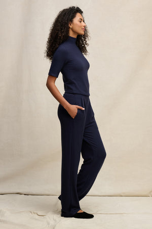A woman with curly hair stands in profile, wearing a navy blue short-sleeve turtleneck and the Velvet by Graham & Spencer ELIYA SWEATPANT—ultra-soft, straight leg sweatpants with her hands in her pockets against a neutral background.