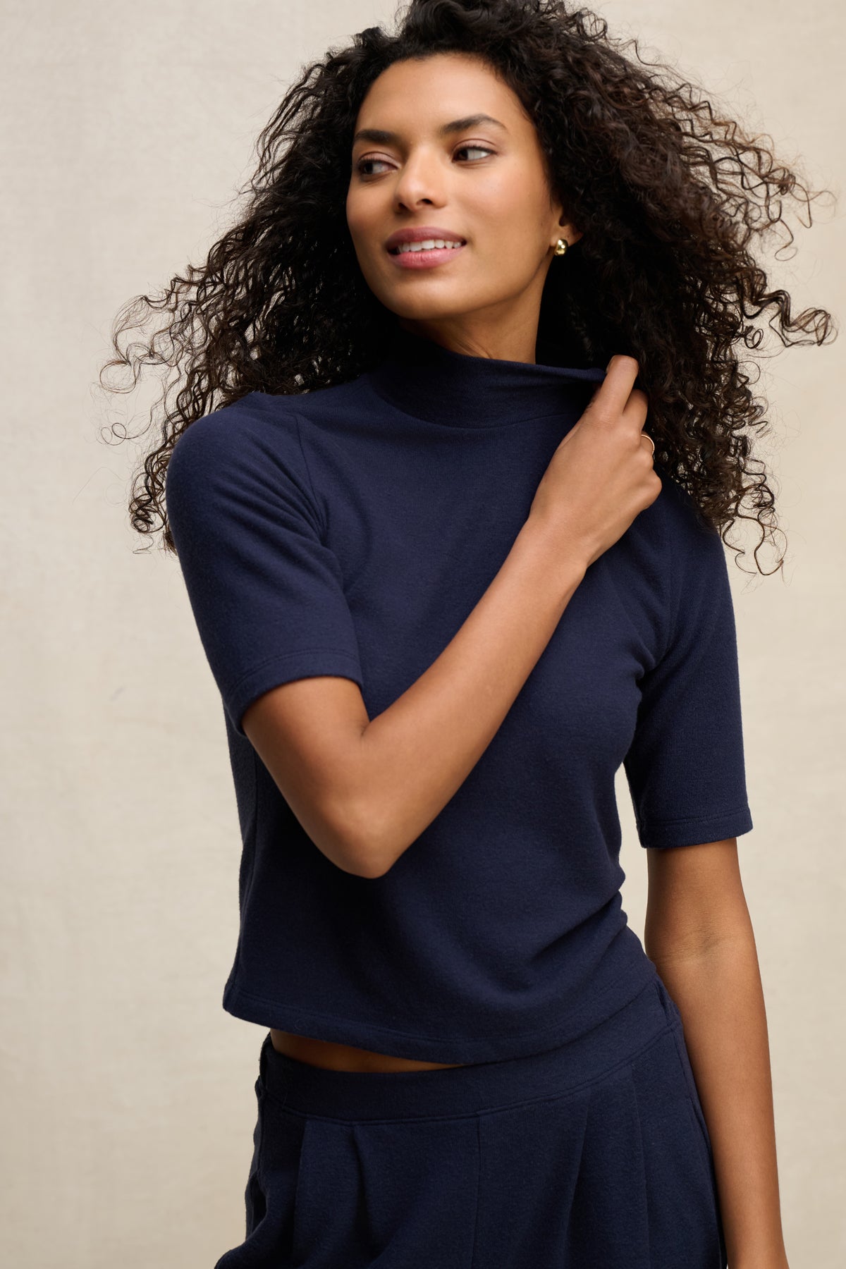 A woman wears the MALAYA TOP by Velvet by Graham & Spencer in navy blue with a mock neck and boxy fit, paired with matching pants, standing against a neutral background and touching her collar.-39838361747649
