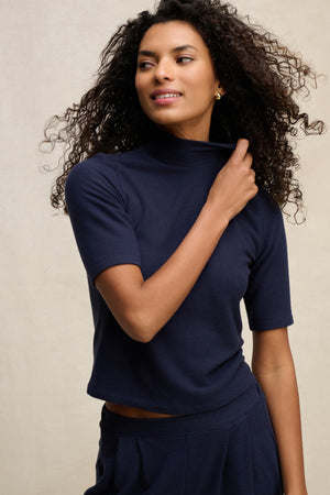 A woman wears the MALAYA TOP by Velvet by Graham & Spencer in navy blue with a mock neck and boxy fit, paired with matching pants, standing against a neutral background and touching her collar.
