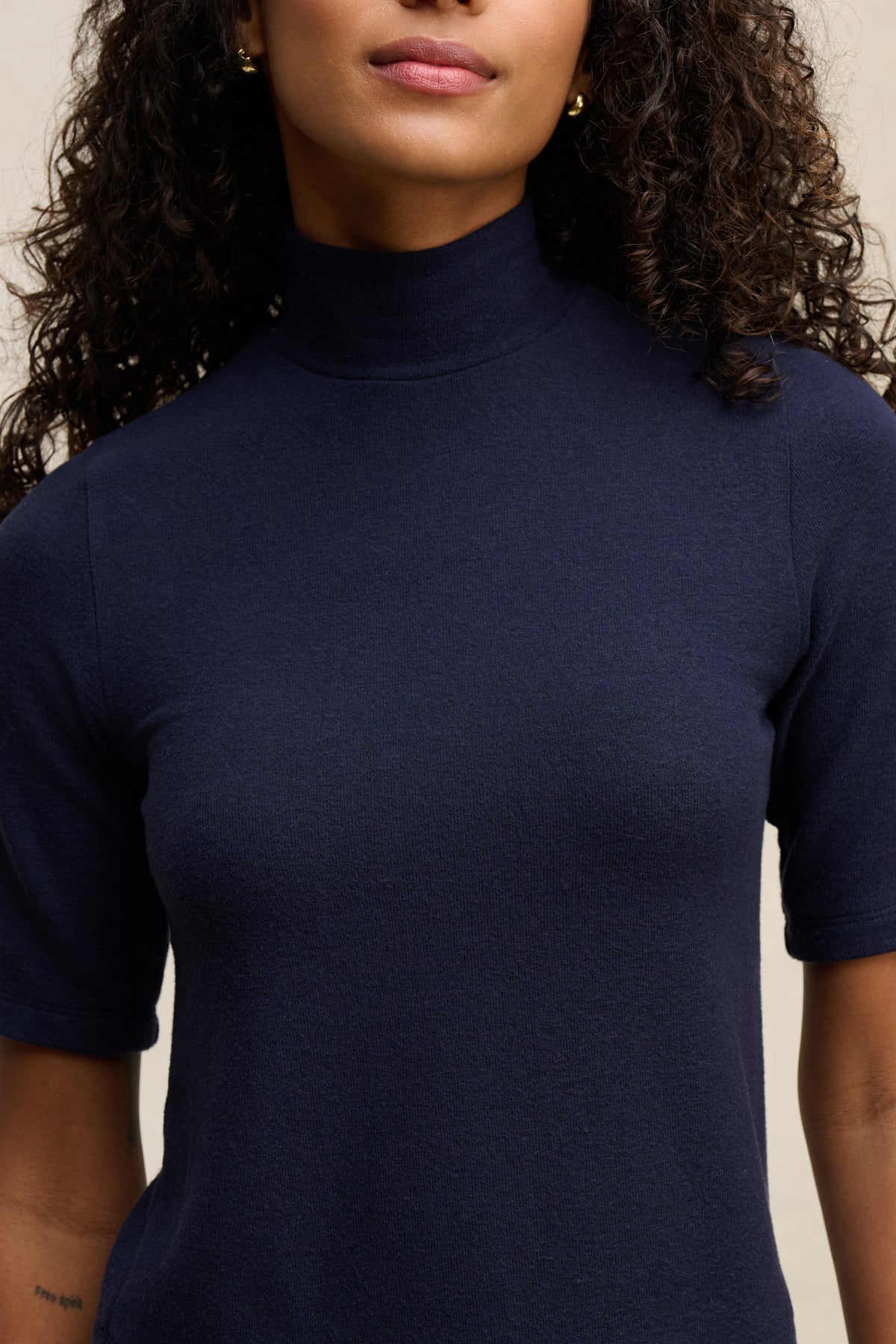 A woman with curly hair models the Velvet by Graham & Spencer MALAYA TOP in navy. Only her lower face and upper torso are shown, emphasizing the boxy fit and short-sleeve mock neck design.-39838361714881