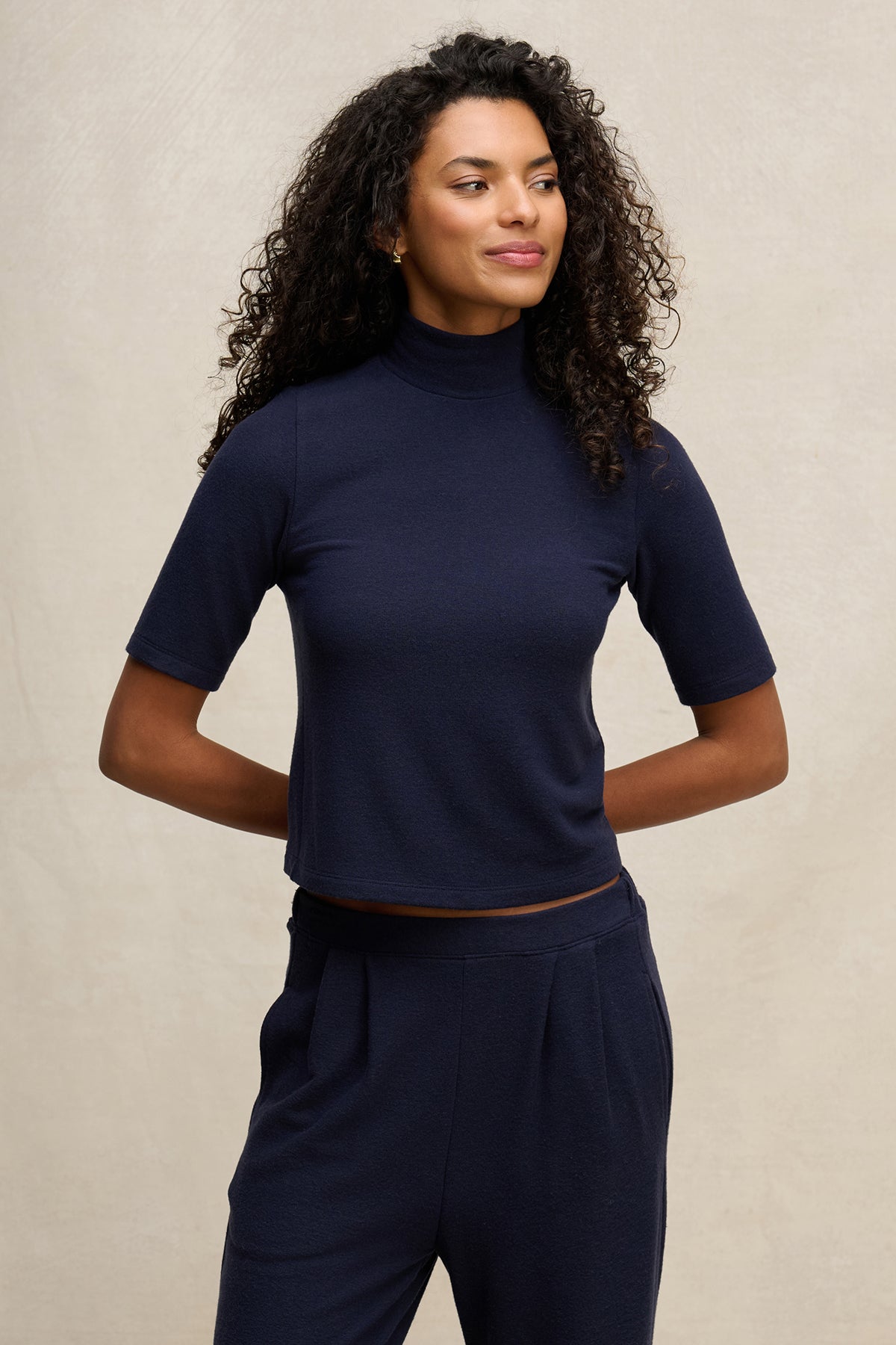 A woman with curly hair wears the MALAYA TOP by Velvet by Graham & Spencer— a navy short-sleeve mock neck boxy fit top—paired with matching pants, standing against a plain light background.-39838361682113