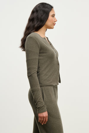 A woman stands in profile wearing the RAYA CARDIGAN by Velvet by Graham & Spencer in olive green against a plain light background.