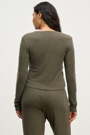 A woman stands with her back to the camera, wearing the RAYA CARDIGAN by Velvet by Graham & Spencer—an olive green, long-sleeve cotton-viscose blend top with matching pants—against a plain background.