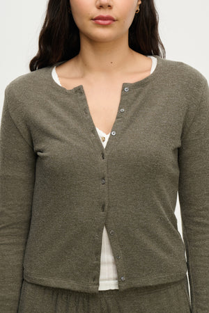 A woman wears the RAYA CARDIGAN by Velvet by Graham & Spencer, an olive green cotton-viscose button front cardigan, over a white top with the top button undone.