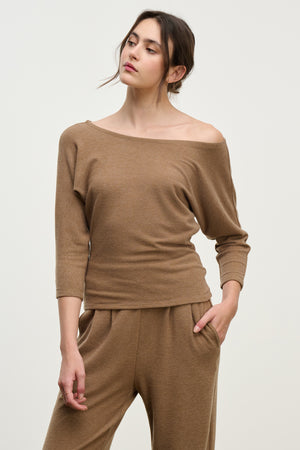 A woman wearing the SELENA TOP by Velvet by Graham & Spencer, a brown cotton viscose boatneck long-sleeve top paired with matching pants, stands against a plain light background with one hand in her pocket.