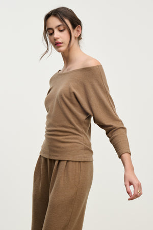 A woman models the SELENA TOP by Velvet by Graham & Spencer, a light brown off-the-shoulder cotton-viscose piece with a relaxed silhouette, paired with matching pants, standing against a plain light background.