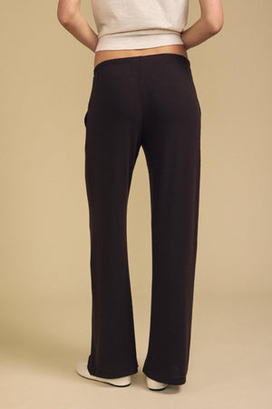 A person stands with their back to the camera, wearing the MACKENZIE LOUNGE PANT by Velvet by Graham & Spencer—a pair of black, wide-leg lounge pants crafted from a soft knit—paired with white shoes and a light-colored top against a beige background.